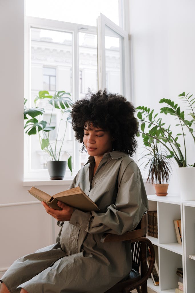 Serene pregnant woman sitting indoors reading a book by a window.